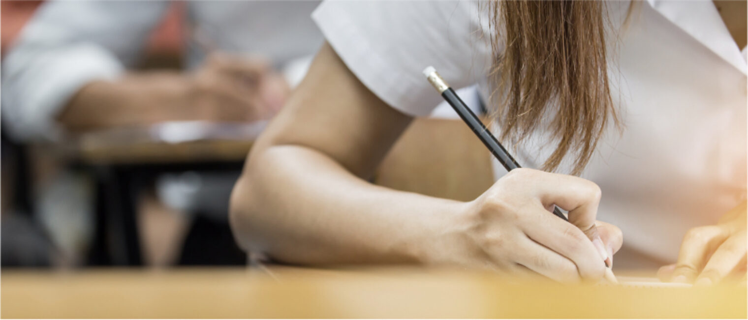 Schriftliche Prüfung: eine Frau im Vordergrund, zu sehen nur ihren Arm mit einem Bleistift, schreibend. Im Hintergrund ein junger Mann mit der gleichen Körperhaltung.