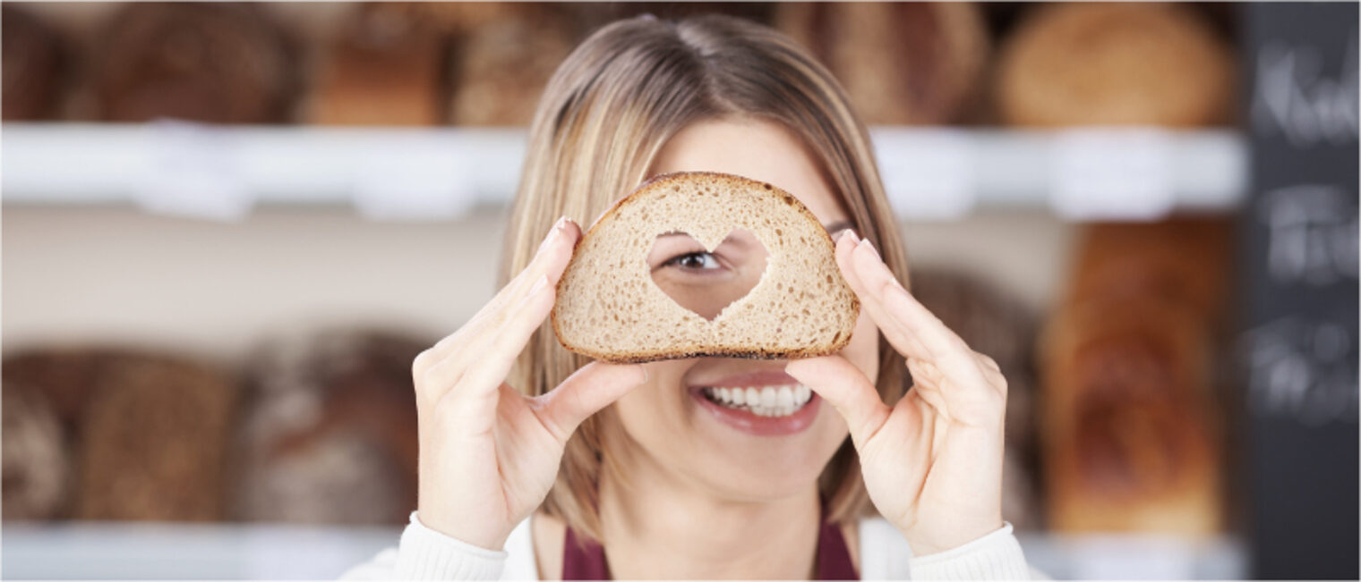 Eine junge Bäckereiverkäuferin die sich lächelnd eine Scheibe Brot, in dem ein Herz ausgeschnitten ist, vor das recht Auge hält.