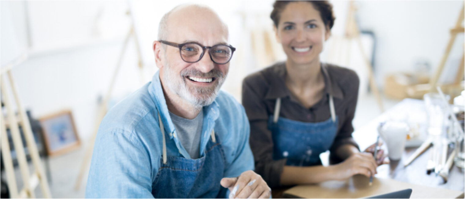 Ein älterer man und eine junge Frau die in einer Werkstatt an einem Tisch stehen und lächelnd in die Kamera schauen.