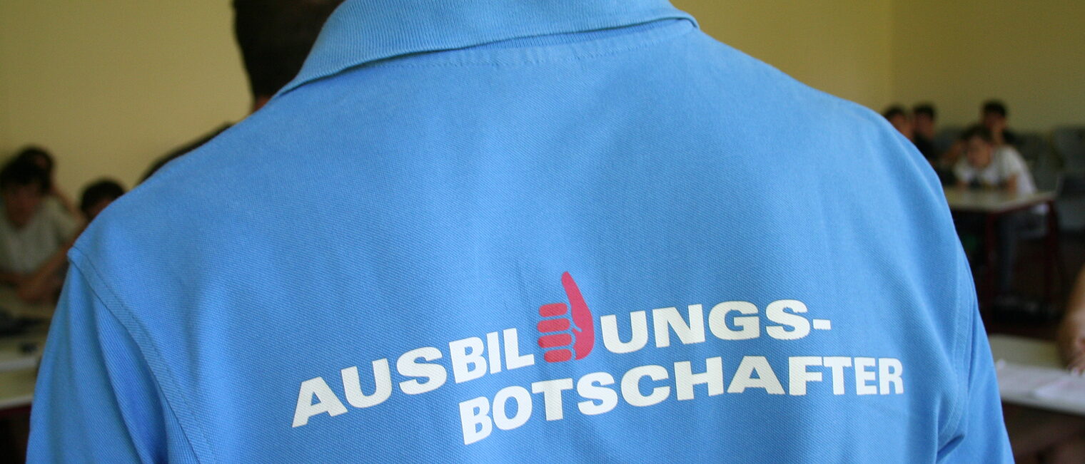 Junger Mann mit blauem T-Shirt mit der Aufschrift "Ausbildungsbotschafter", der vor einer Schulklasse spricht.