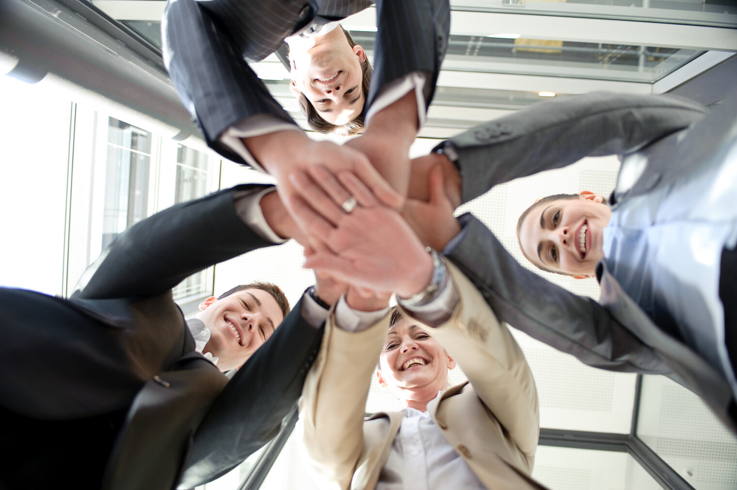 Fotoperspektive von unten: vier Personen im Businessoutfit laecheln und legen die Haende in der Mitte uebereinander.