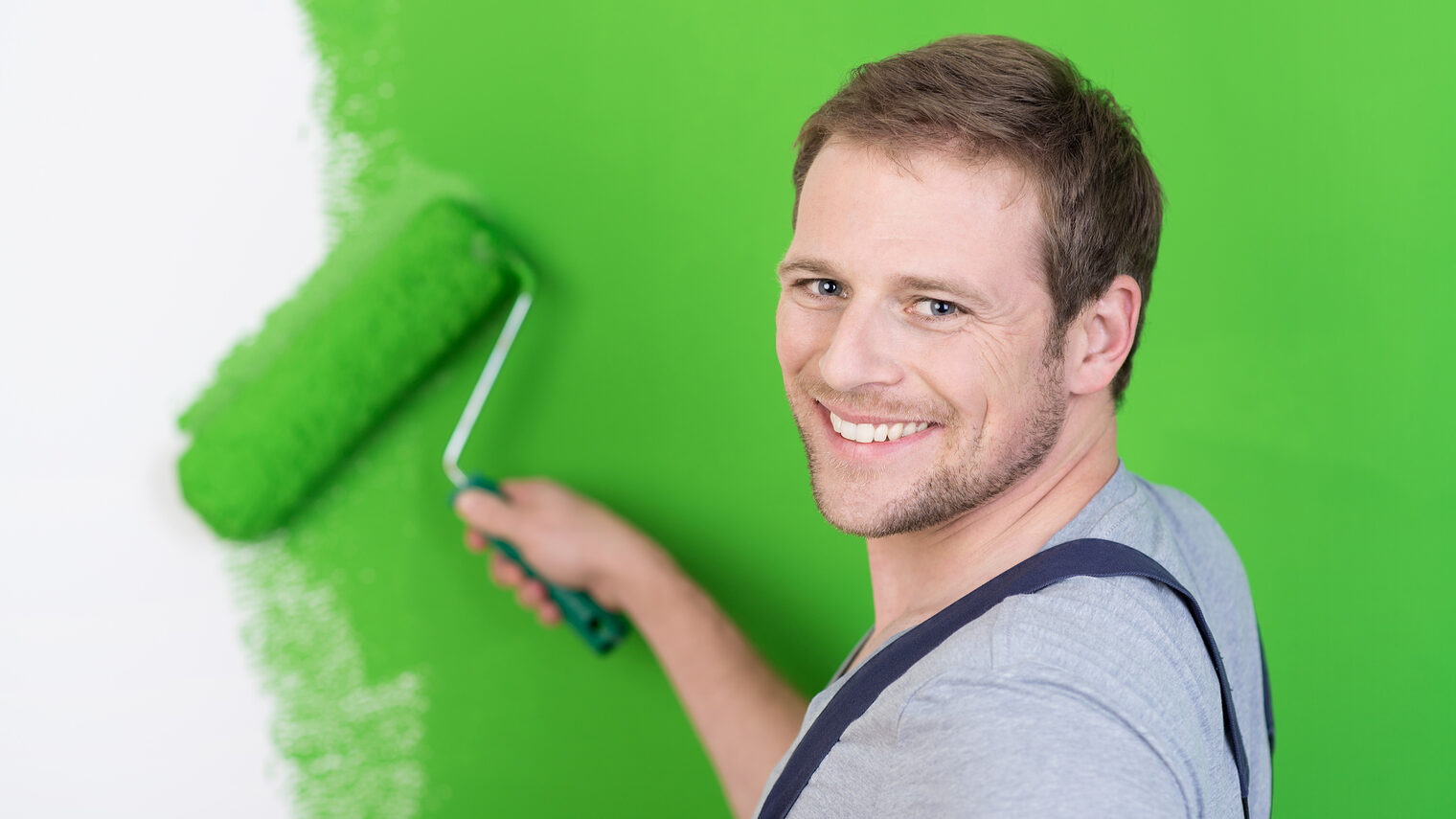 Ein Mann mit Latzhose streicht mit einer Rolle in der Farbe grün eine Wand an. 