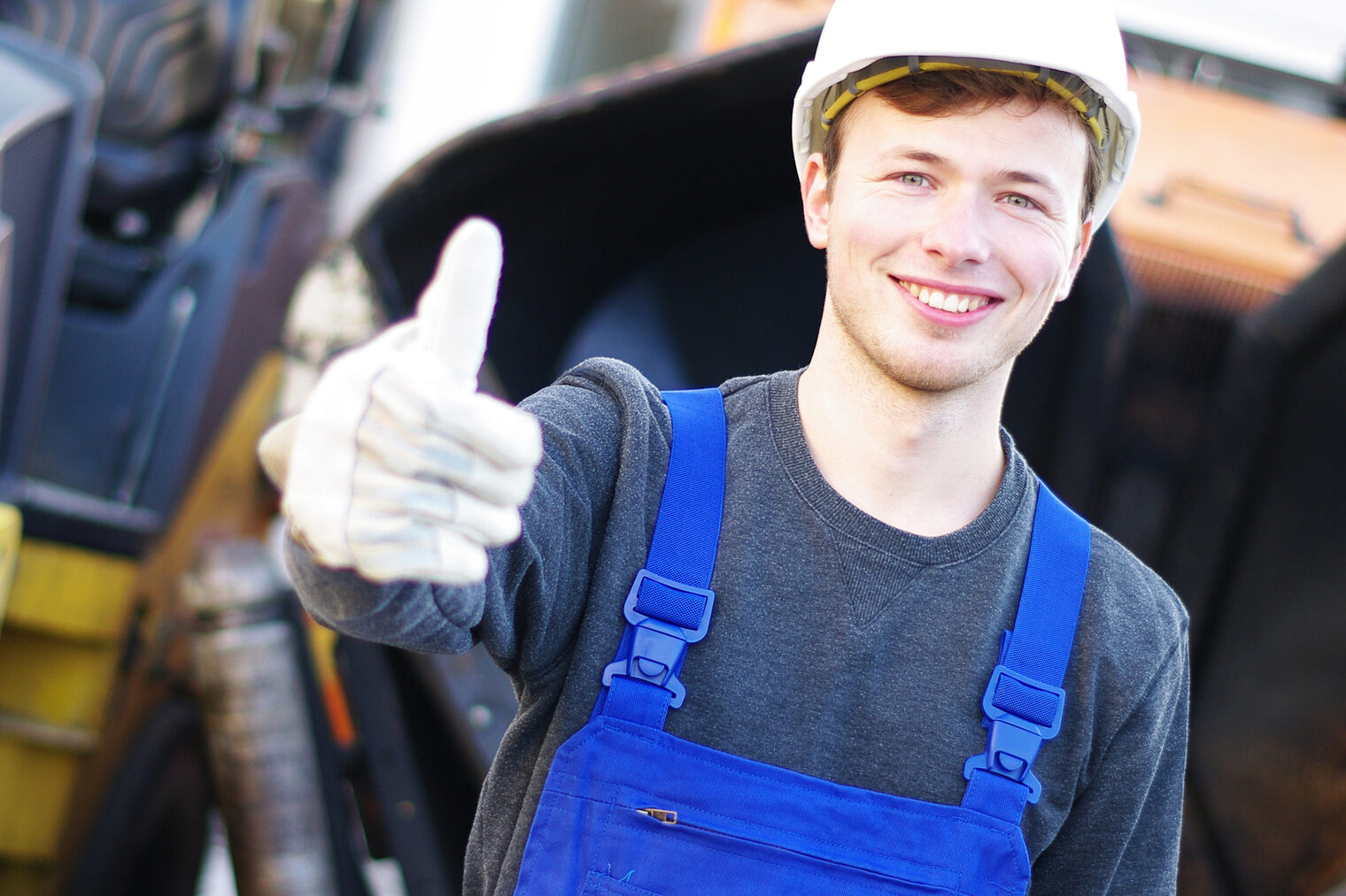Junger Mann mit weißem Schutzhelm auf dem Kopf, blauer Latzhose und weißen Handschuhen. Im Hintergrund ein gelbes Baufahrzeug, hebt den Daumen in die Höhe.