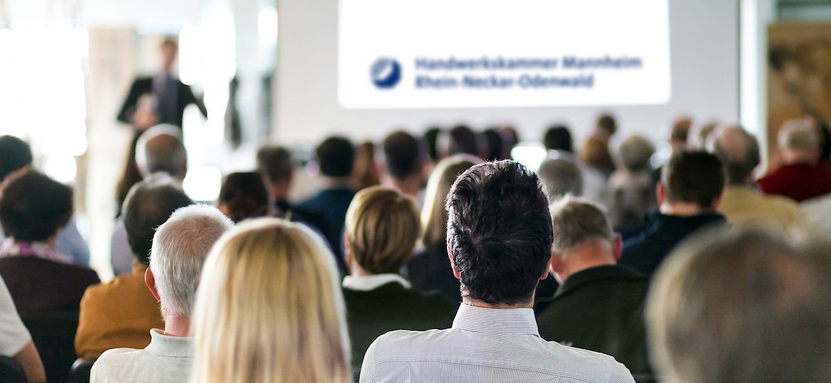 Speaker Giving a Talk at Business Meeting. Audience in the conference hall. Business and Entrepreneurship. Panoramic composition suitable for banners. Schlagwort(e): event, lecture, business, speech, meeting, talk, speaker, seminar, lecture hall, conference, convention center, workshop, entrepreneur, educator, education, explaining, listeners, participants, group, training, professional, corporate, businessman, coaching, briefing, manager, man, academy, faculty, congress, summit, institute, podium, knowledge, screen, university, presentation, public, people, sitting, study, audience, academic, convention, information, symposium, discussion, entrepreneurship, expert, teach