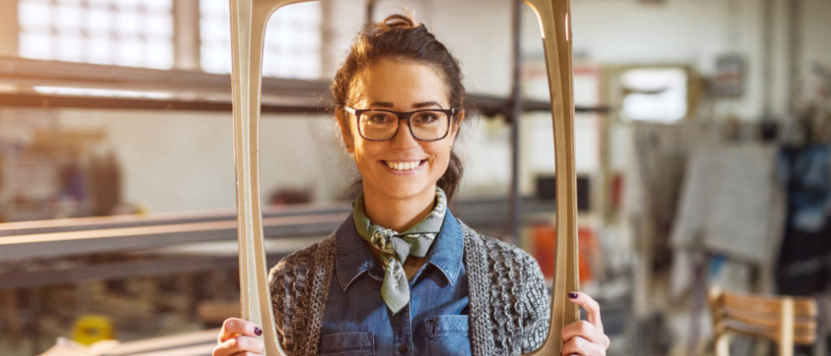 Excited middle-aged engineer holding old fashion picture frame in sunny fabric workshop and posing behind.