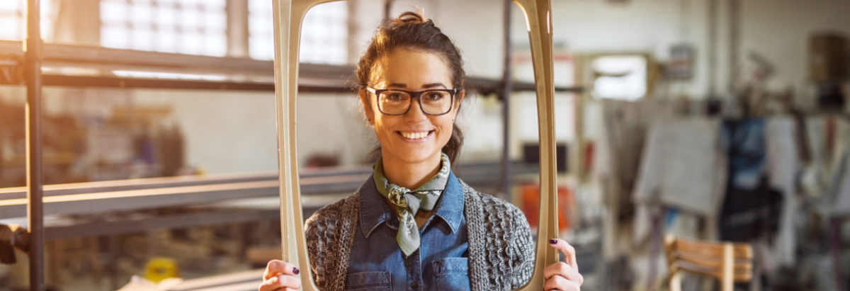 Excited middle-aged engineer holding old fashion picture frame in sunny fabric workshop and posing behind.