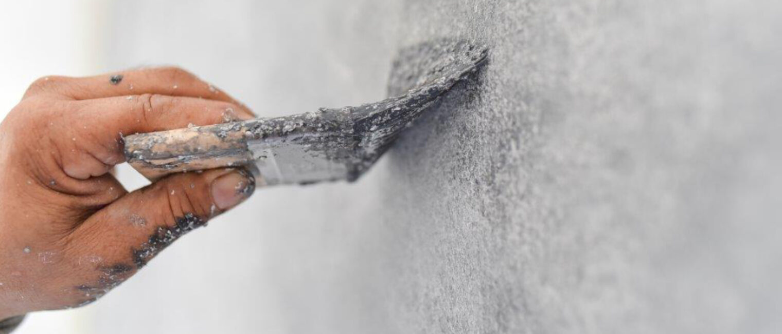 Stuckateur verteilt Putz an der Wand. Zu sehen ist eine Hand mit einem Verteilerpinsel in der Hand. 