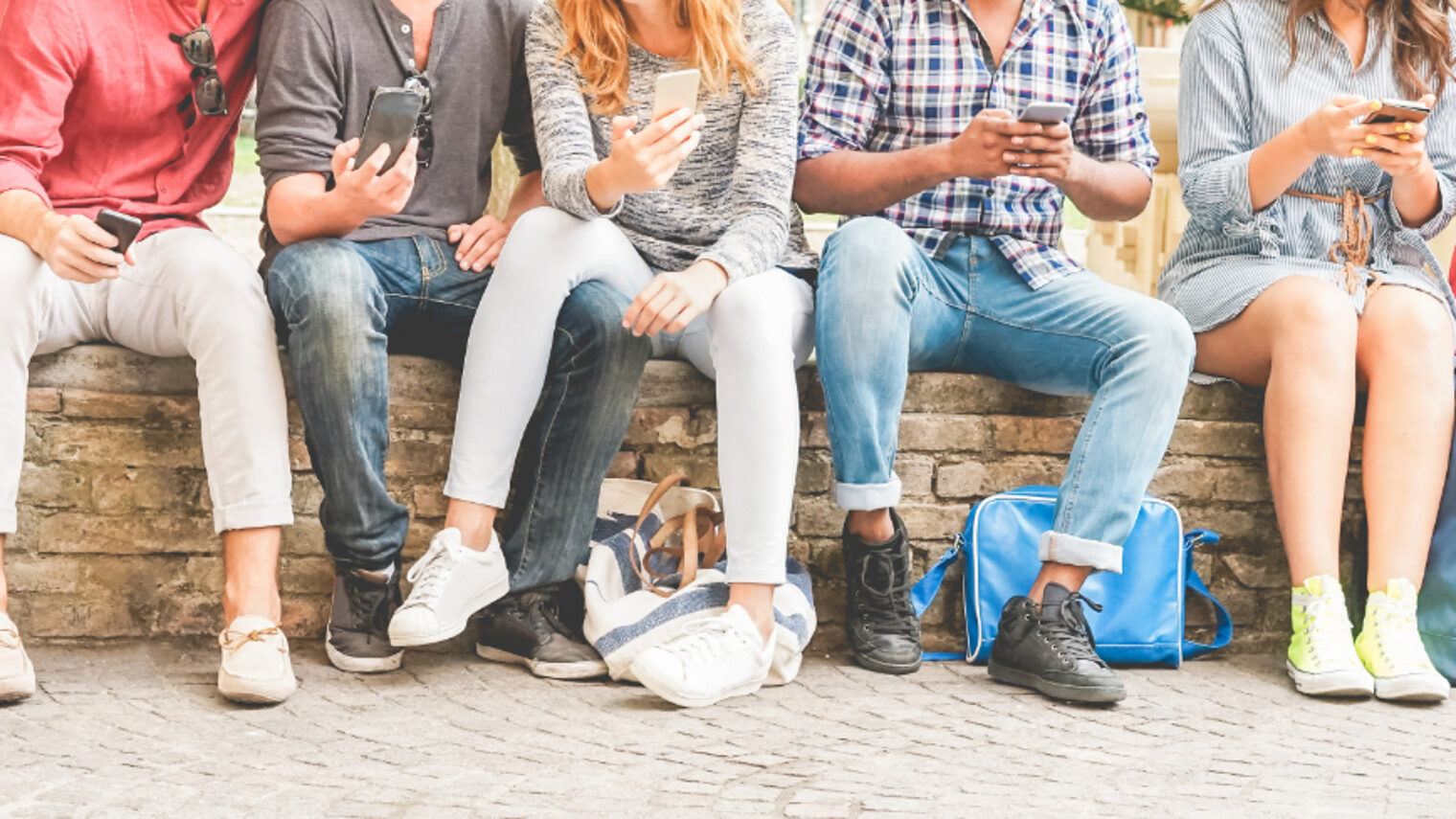 Sieben Jugendliche sitzen auf einer Mauer und spielen am Handy. Die Gesichter sind nicht zu sehen