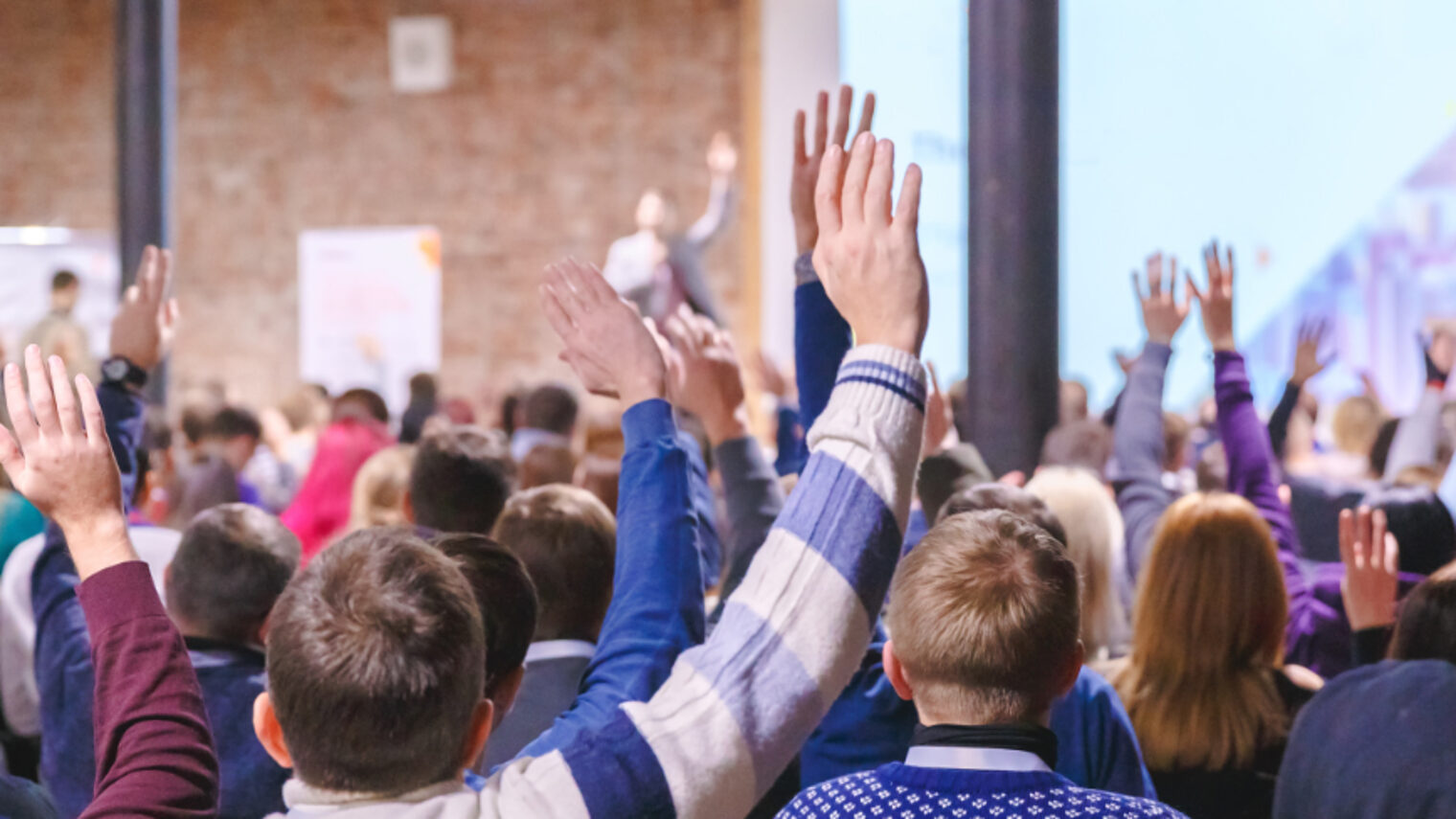 Im Hintergrund steht ein Redner auf einer Bühne; im Vordergrund sitzen Schüler und Schülerinnen von hinten fotografiert. Mehrere Personen heben die Hand für eine Wortmeldung.