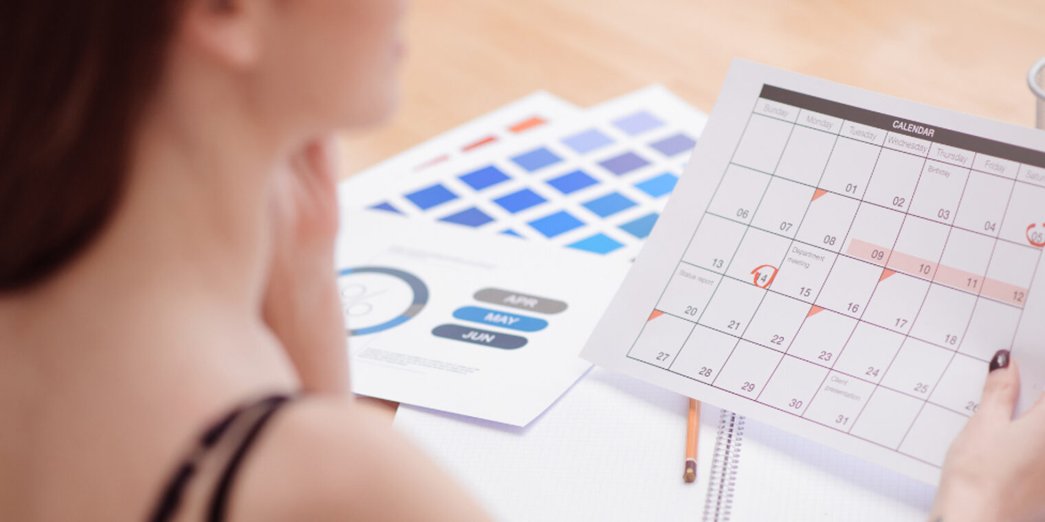Eine Frau hält einen Kalender vor sich, bei dem einige Tage rot umkreist sind. Im Hintergrund liegen weitere Unterlagen.