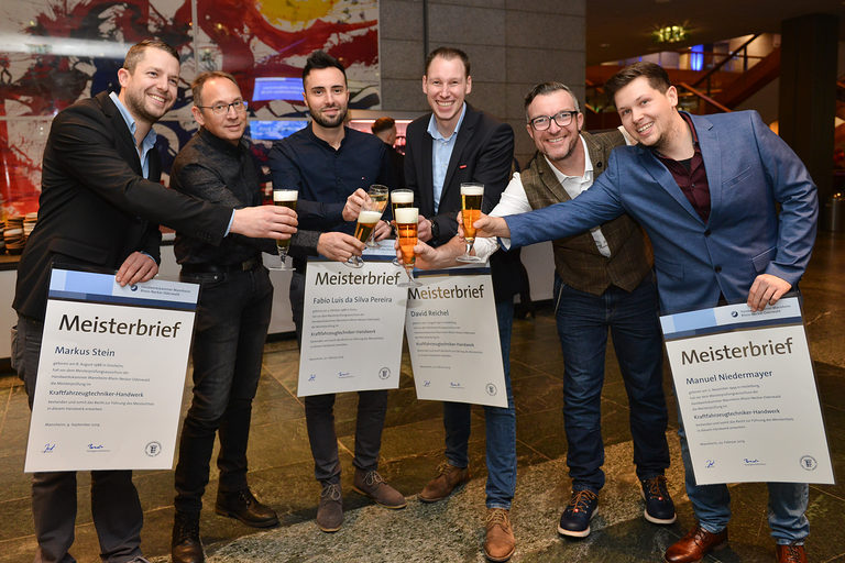 Sechs Meister halten ihren Meisterbrief in der Hand uns stoßen mit einem Bier darauf an. 