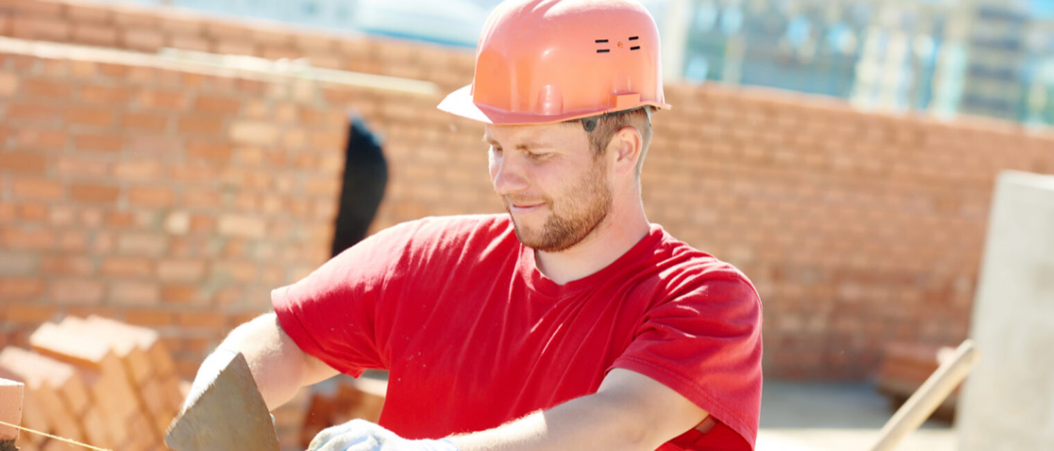Bauarbeiter mit rotem T-Shirt und Helm in der Farbe orange zieht eine Mauer hoch