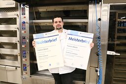 Bäcker- und Konditormeister zeigt seine Meisterbriefe vor seinem Backofen.