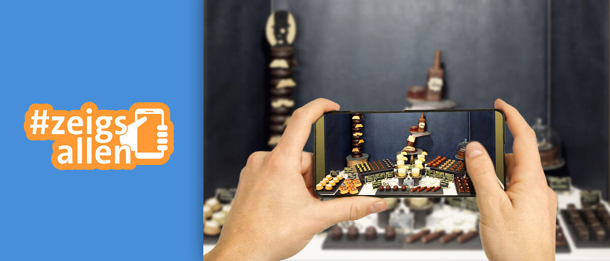 Fotowettbewerb #zeigsallen Logo des Fotowettbewerbs #zeigsallen und zwei Hände, die mit einem Handy einen dekorierten Tisch einer Konditoren-Meisterprüfung fotografieren.