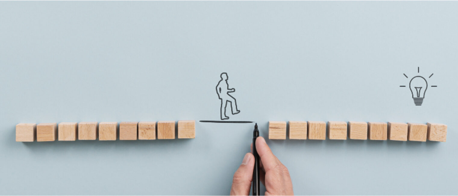 Holzwürfel auf einem Blatt; in der Mitte eine größere Lücke; über die Lücke zeichnet eine Hand eine Linie und darauf stehenden eine Person, am Ende ist eine Glühbirne gezeichnet.