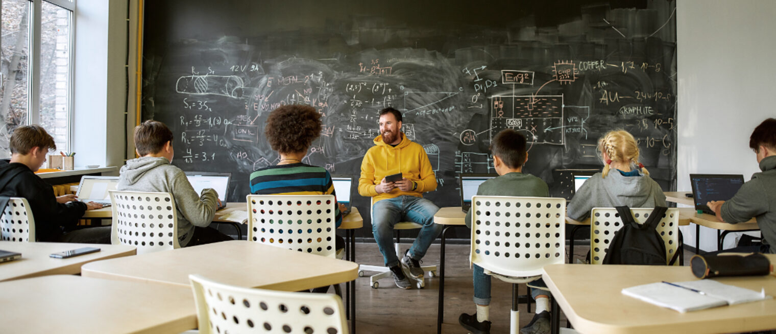 Ein Lehrer sitzt von einer vollgeschriebenen Tafel und um ihn herum sechs Schüler.