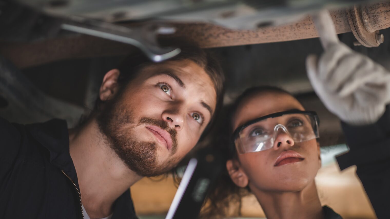 Ein Kraftfahrzeugtechniker und eine Kraftfahrzeugtechnikerin prüfen die Unterseite eines Autos. 