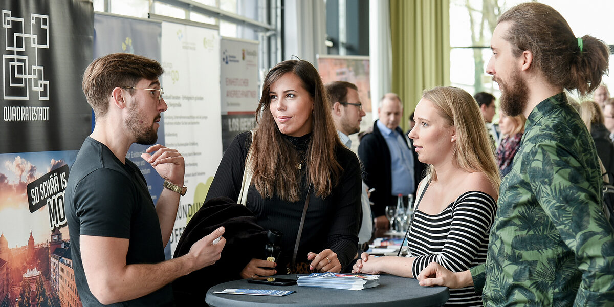 Start Rhein-Neckar Zwei Frauen und zwei Männer stehen auf einer Messe an einem rundne Stehtisch zusammen und erzählen