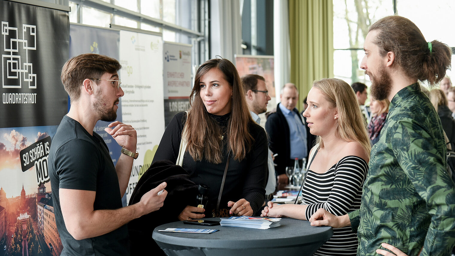 Zwei Frauen und zwei Männer stehen auf einer Messe an einem rundne Stehtisch zusammen und erzählen