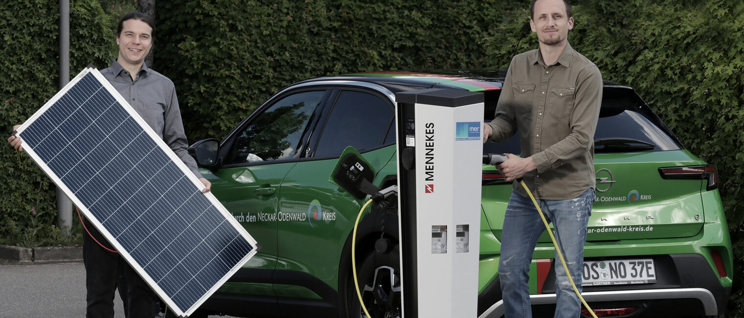 Zwei Männer, einer hält ein Solar-Panel in der Hand, der Andere steht an einer Ladesäule mit der Hand am Ladestecker. Bei vor einem grünen Kleinwagen.