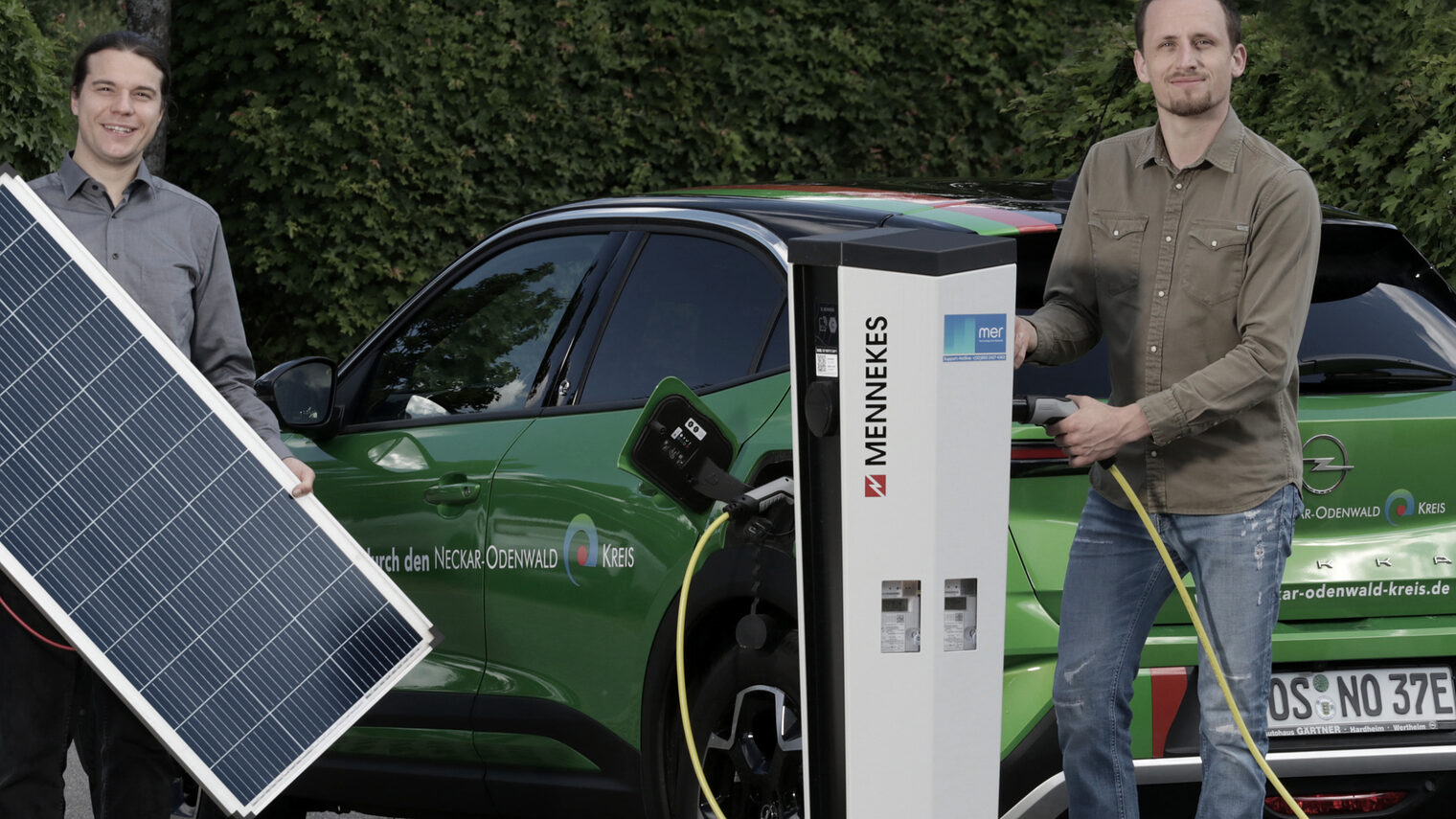 Zwei Männer, einer hält ein Solar-Panel in der Hand, der Andere steht an einer Ladesäule mit der Hand am Ladestecker. Bei vor einem grünen Kleinwagen.
