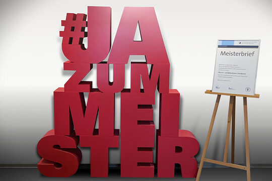 Schriftzug "#JAzumMEISTER" in 3D-Optik neben einem Meisterbrief auf einer Staffelei