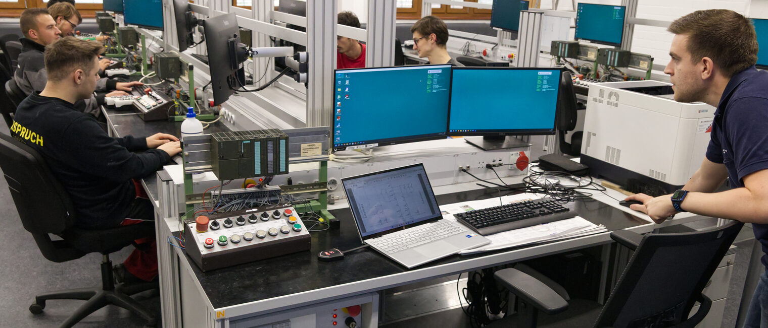 Werkstatt Schreibtische mit Laptop, Bildschirme, Schaltpulte. Vor den Schreibtischen sitzen junge Menschen auf Rollstühlen.