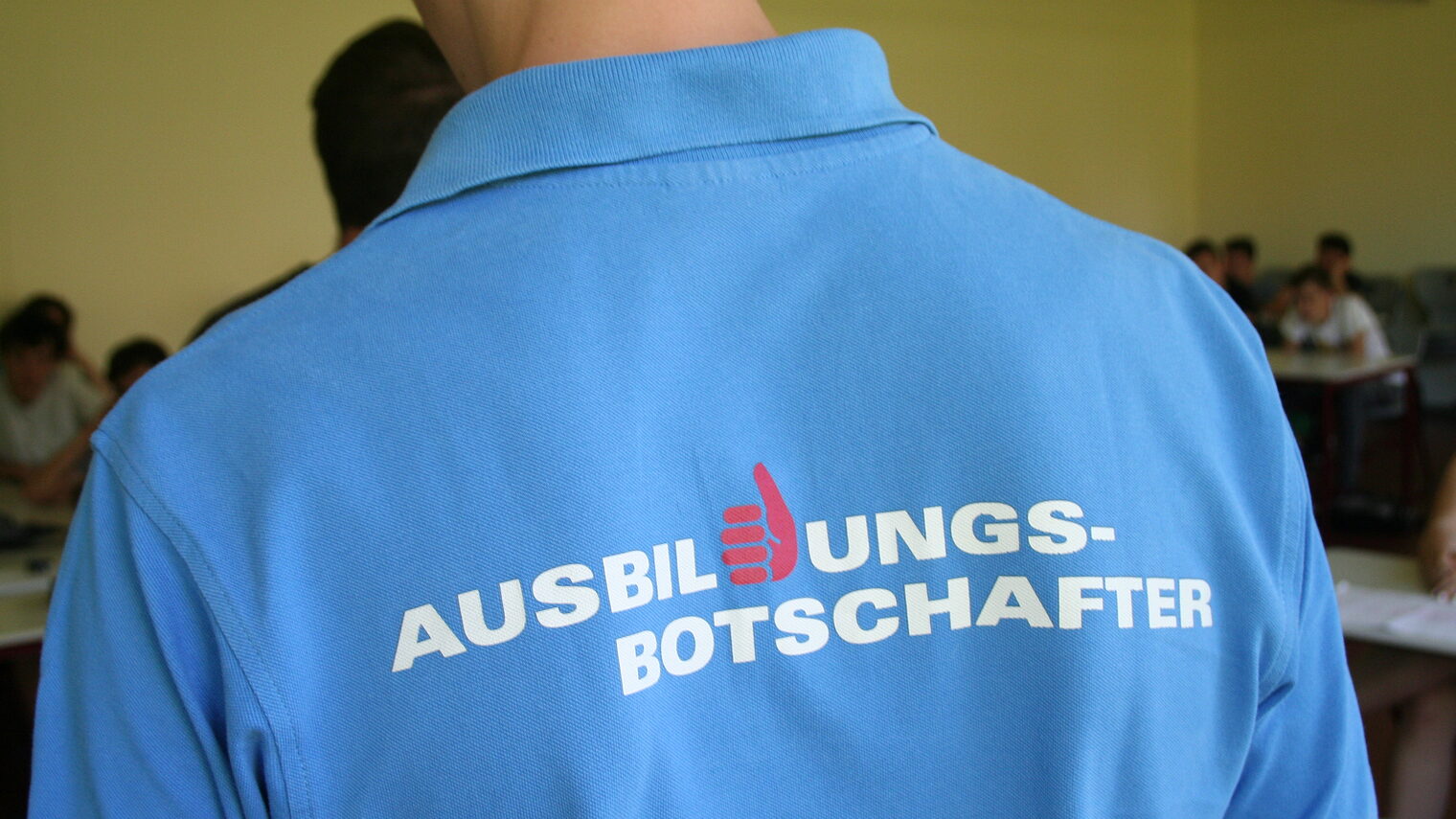 Junger Mann mit blauem T-Shirt mit der Aufschrift "Ausbildungsbotschafter", der vor einer Schulklasse spricht.