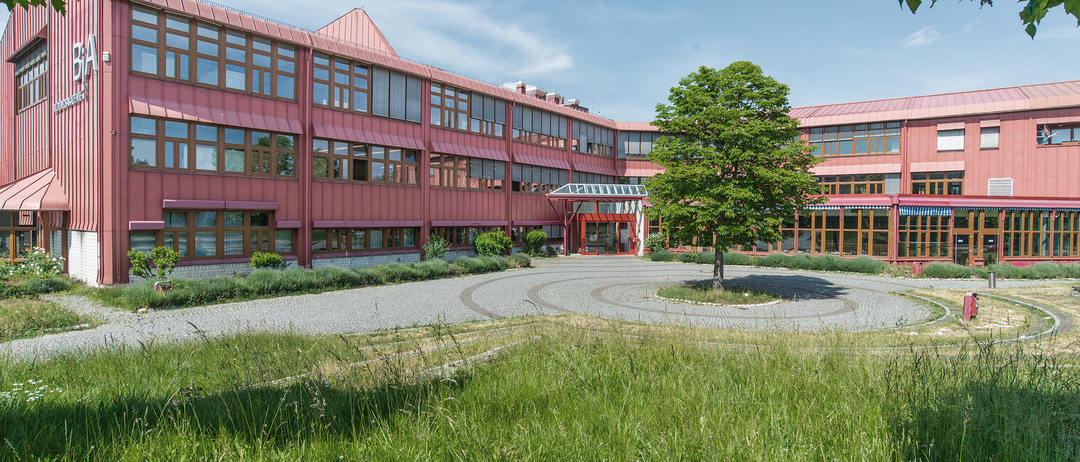 Die Bildungsakademie der Handwerkskammer ist ein rotes Gebäude mit großen Fensterfronten, davor ein Baum und grüne Wiese.