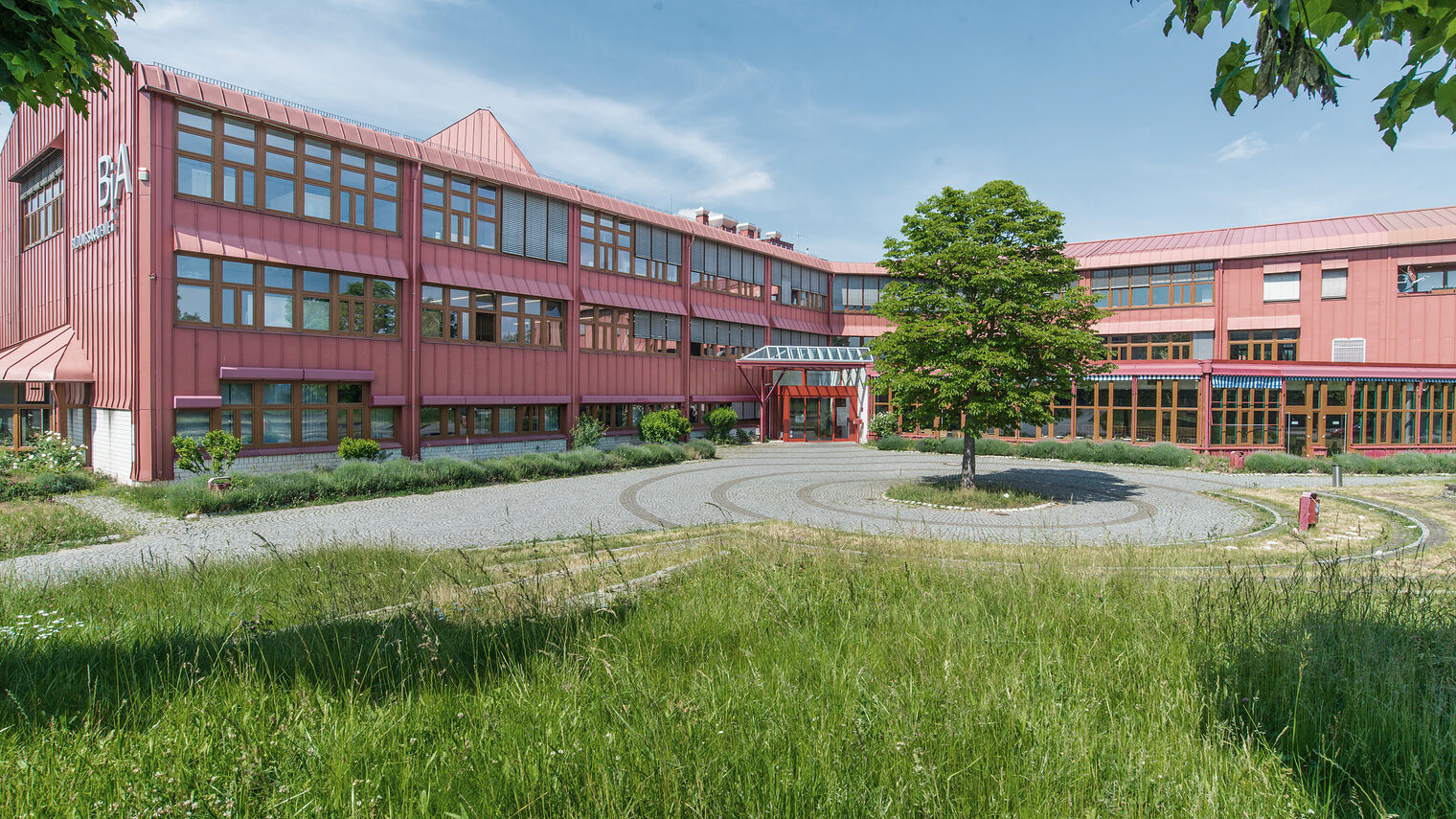 Die Bildungsakademie der Handwerkskammer ist ein rotes Gebäude mit großen Fensterfronten, davor ein Baum und grüne Wiese.