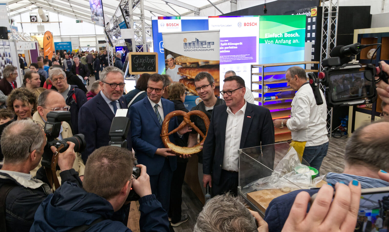 Viele Leute im Zelt. Obermeister hält Riesenbrezel in der Hand. 