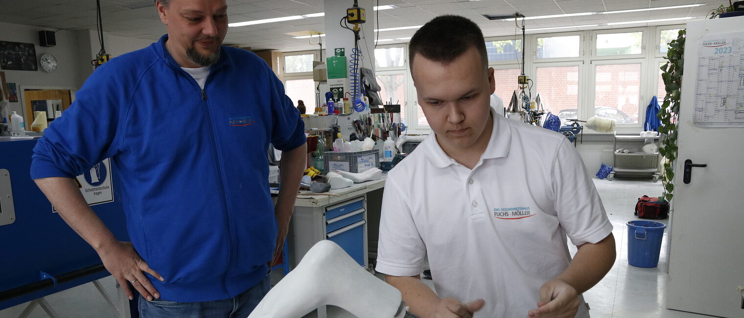 Ausbilder Sebastian Hannen mit blauer Jacke schaut einem Azubi mit weißem Shirt bei der Arbeit zu. 