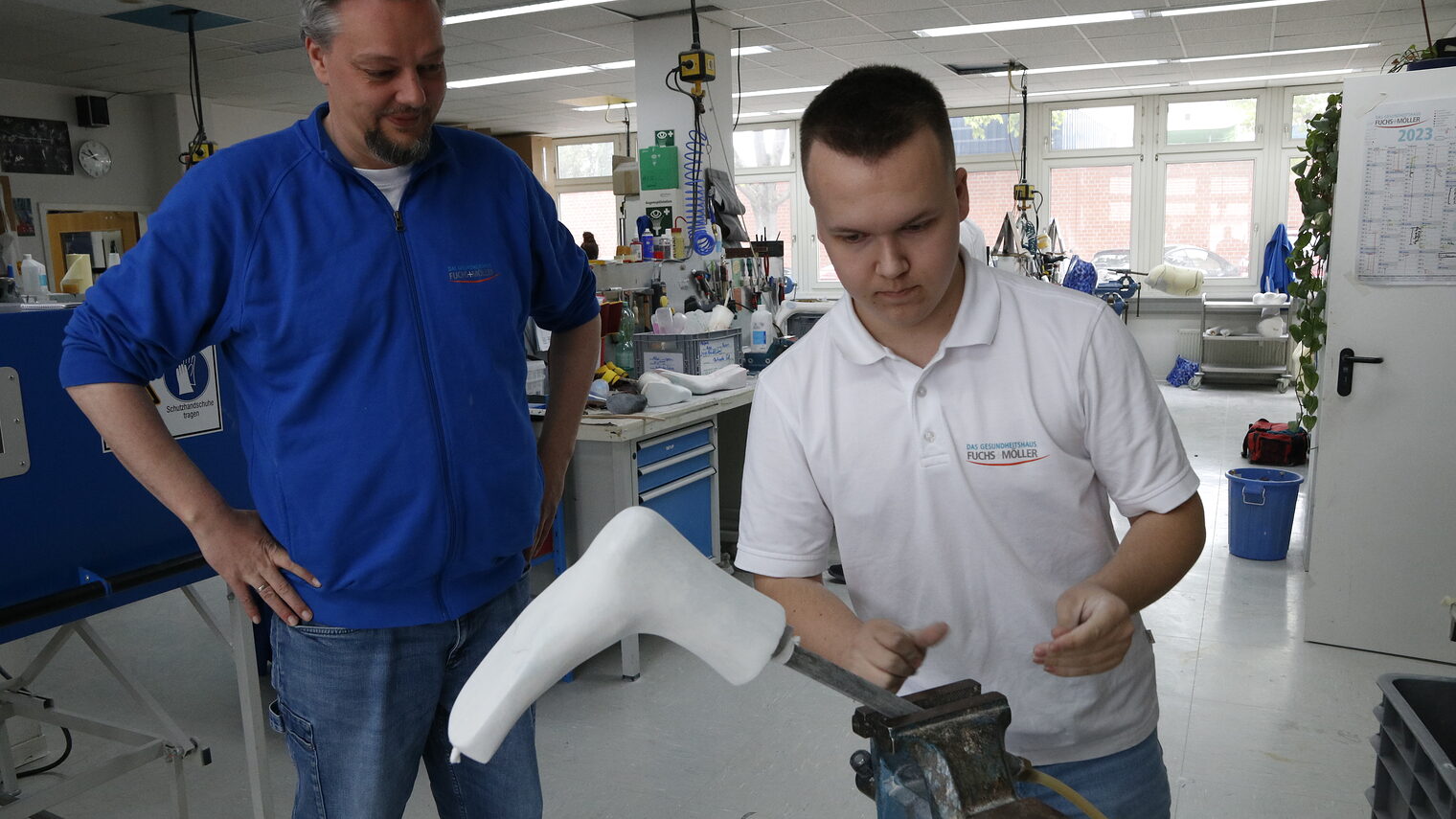 Ausbilder Sebastian Hannen mit blauer Jacke schaut einem Azubi mit weißem Shirt bei der Arbeit zu. 