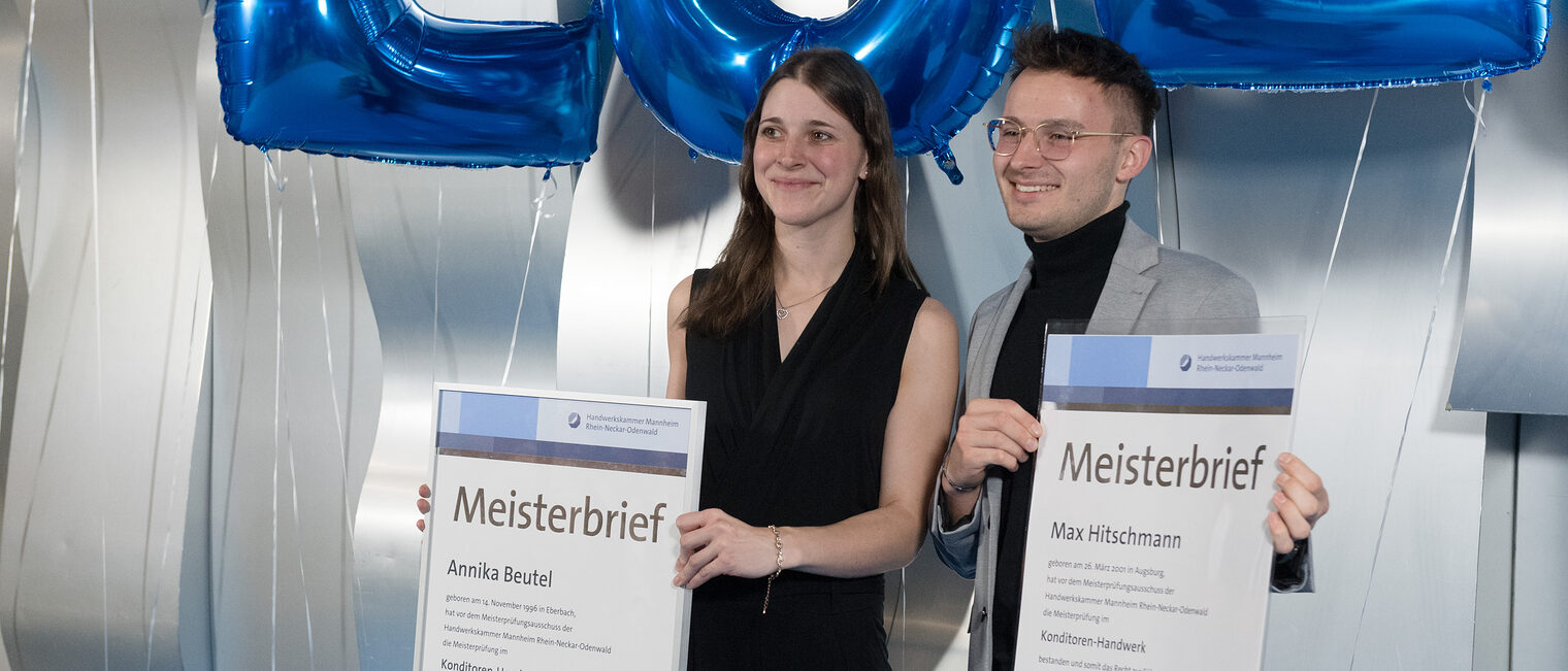 EIne Konditormeisterin und ein Konditormeister lassen sich mit ihren Meisterbriefen in der Hand vor einer Fotowand fotografieren. 