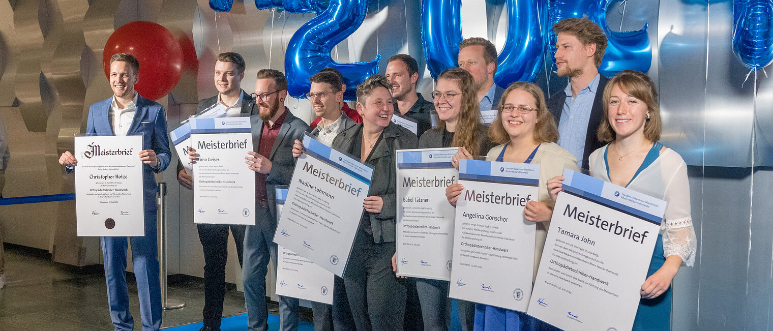 Eine Gruppe Orthopädietechnikermeisterinnen und -meister lässt sich vor einer Fotowand mit ohren Meisterbriefen in der Hand ablichten. 