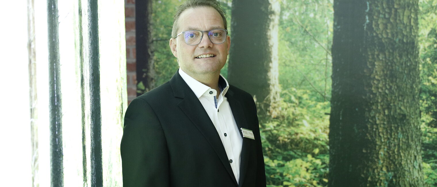 Geschäftsführer Thomas Frey von Koob Hörgeräte steht vor einem Wald den er in seiner Hör-Erlebnis Welt künstlich erschaffen hat.
