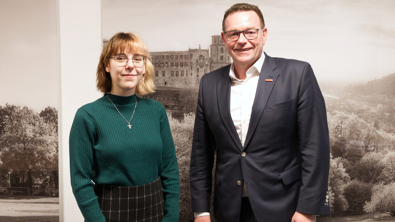 Frau Bette in grünem Pullover mit kariertem Rock rechts stellt sich mit Präsident Hofmann im Anzug links dem Fotografen. 