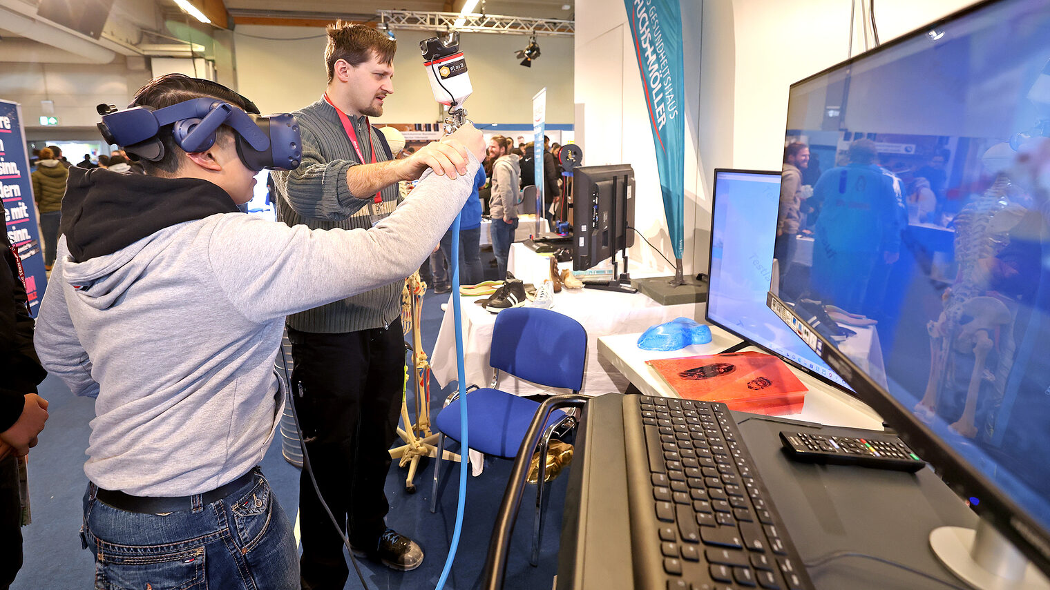 Eine junge hat eine VR-Brille auf und wird von einem jungen Mann angeleitet, diese fachmännisch anzuwenden. 