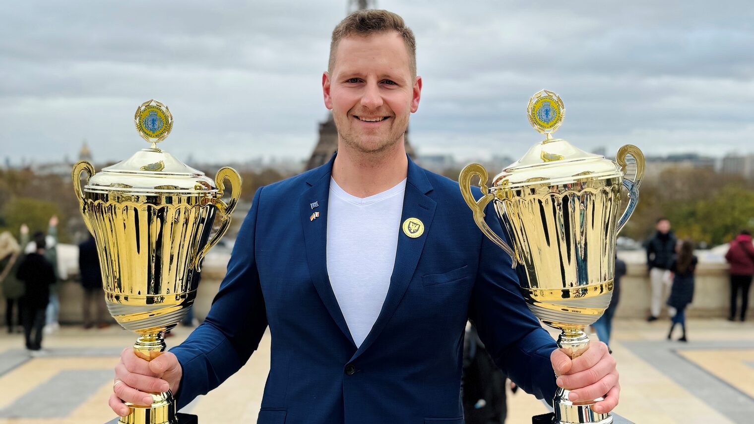 Mann mit weißem Shirt und blauem Jacket hält jeweils einen Pokal in der Hand. 