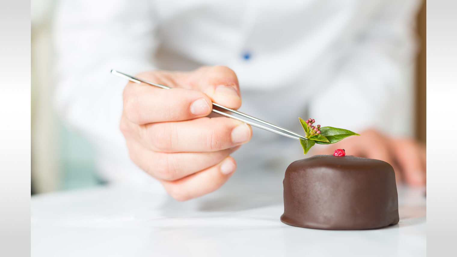EIn Konditor verziert eine kleine Schokoladentorte mit einer Pinzette für Fondant-Deko