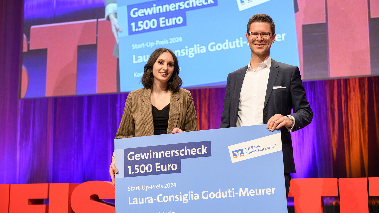 Dunkelhaarige Frau hält zusammen mit einem Mann in weißem Hemd und dunklem Jackett einen großen Scheck der VR Bank in den Händen 