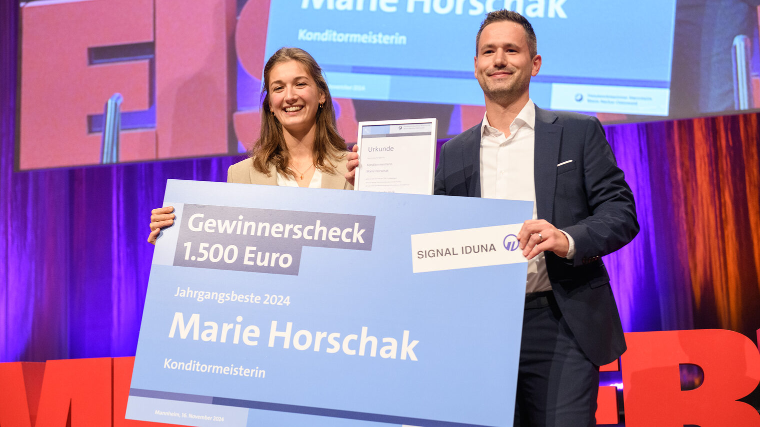 Vertreter einer Versicherung überreicht bei der Meisterfeier einen überdimensionierten großen Scheck an eine Konditormeisterin