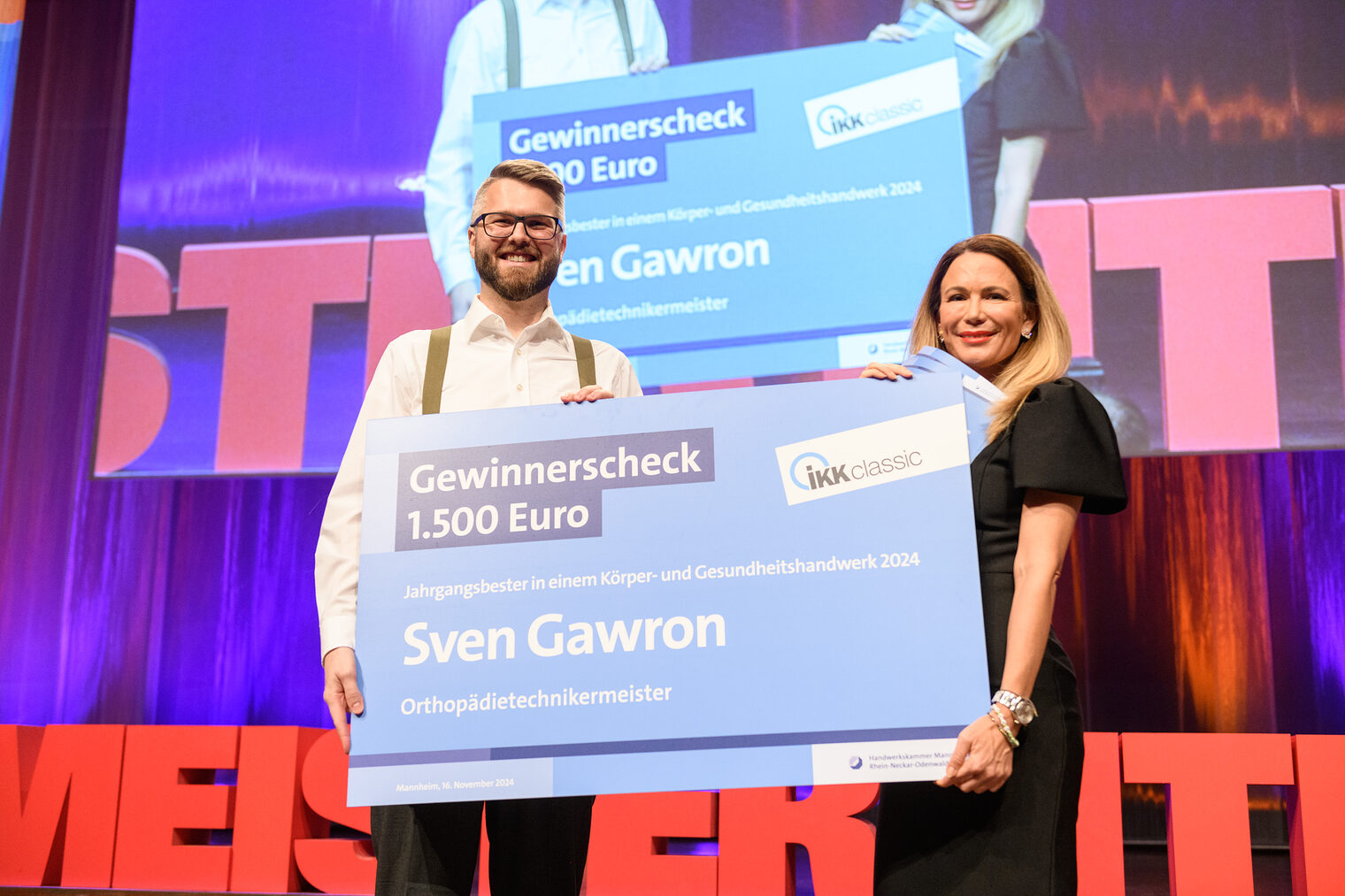 Moderatorin rechts überreicht großen, blauen Scheck an Orthopädietechnikermeister bei der Meisterfeier über 1.500 Euro