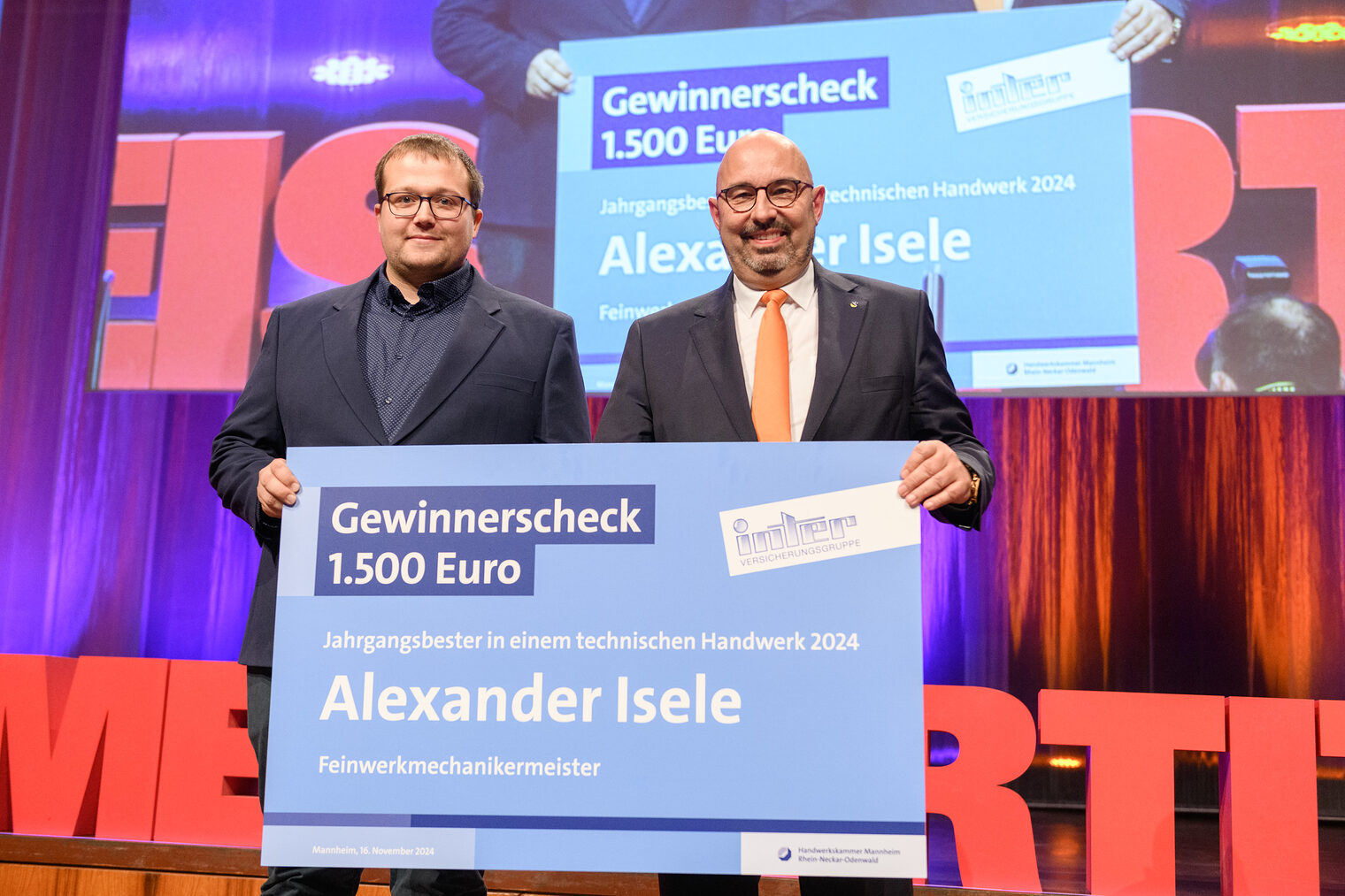 Zwei Männer mit Brille halten einen überdimensioniert großen blauen Scheck über 1.500 Euro vor sich und lachen in die Kamera