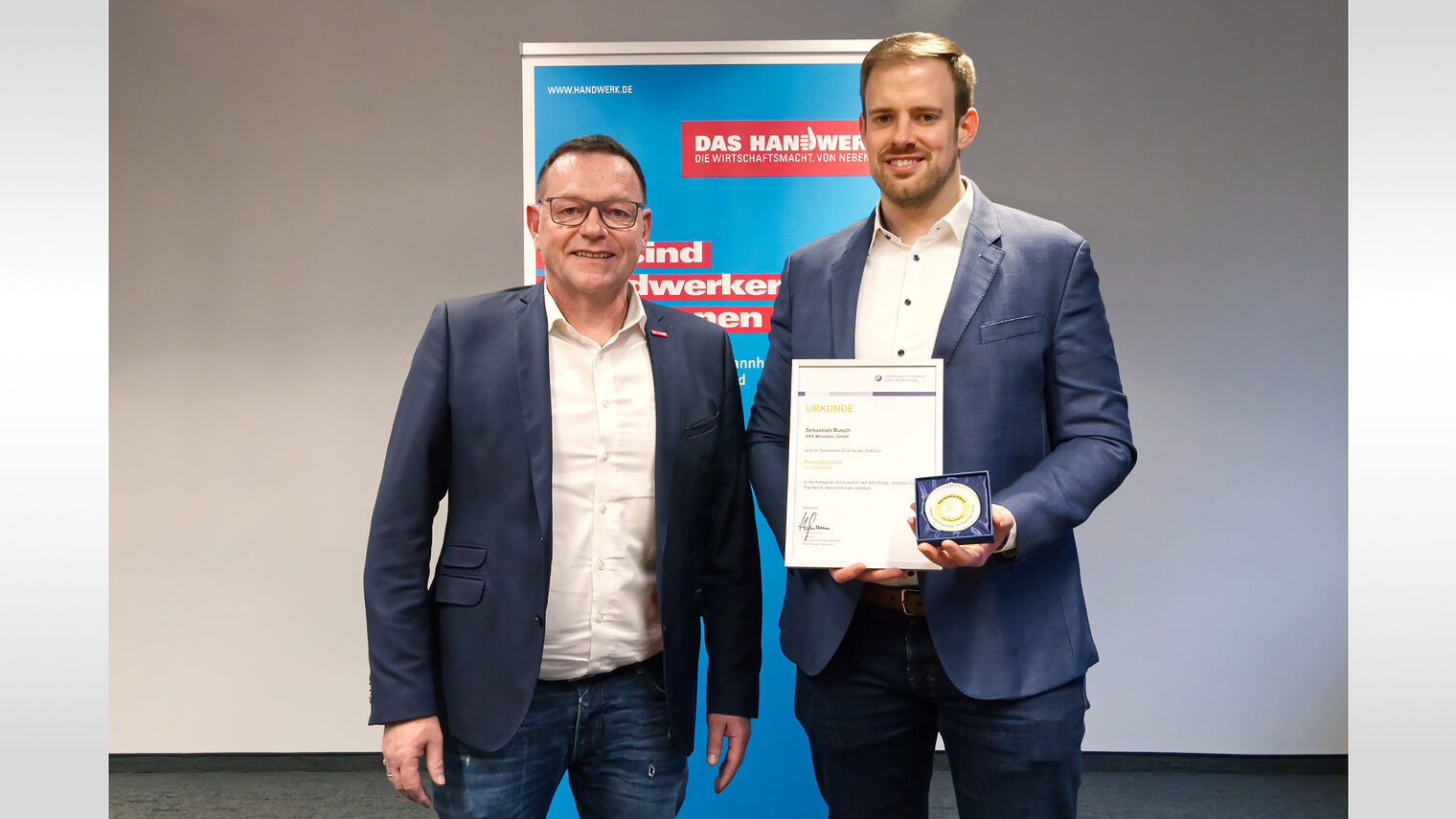 Sebastian Busch stellt sich mit Klaus Hofmann vor einer Plakatwand mit Urkunde und Medaille dem Fotografen. 