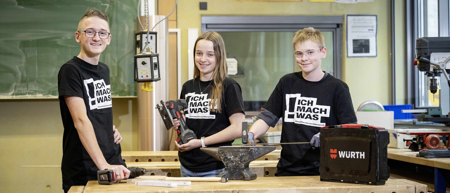 Drei Teenager am Werktisch in einer Schul-Werkstatt, die T-Shirts mit der Aufschrift "Mach was!" tragen und Werkzeuge betätigen.