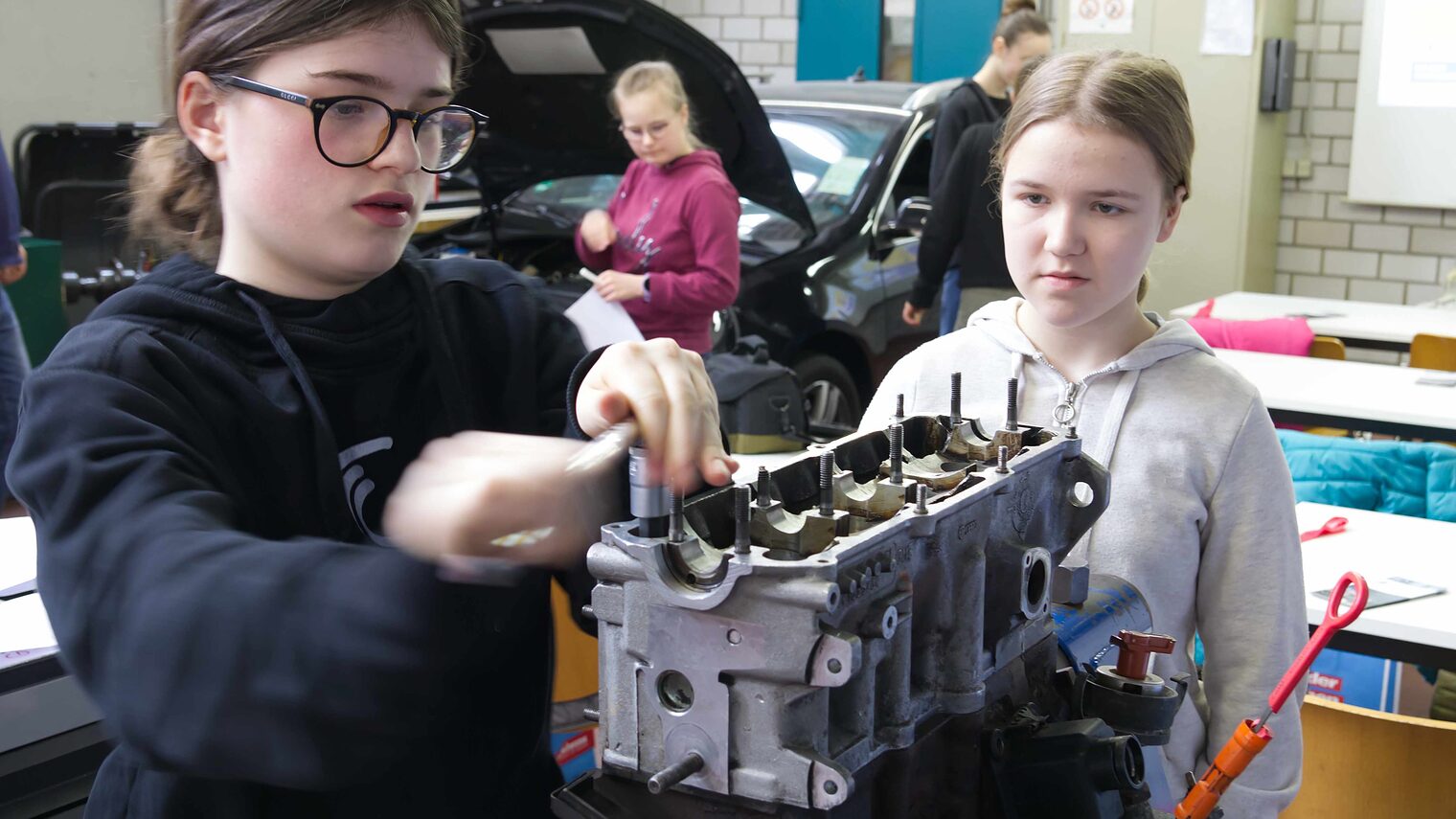 Ein Mädchen mit Brille schraubt in einer Autowerkstatt an einer Batterie. Ein zweites Mädchen beobachtet sie dabei aufmerksam.