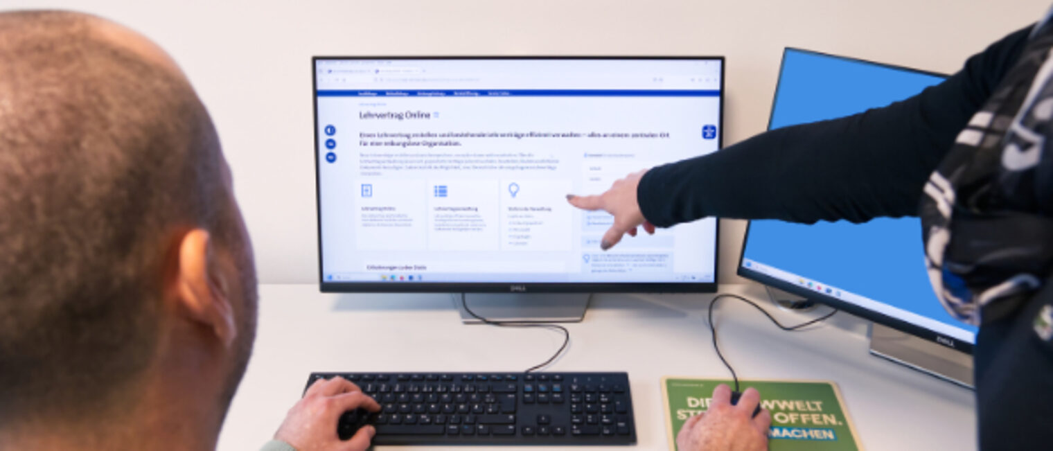 Frau zeigt einem Mann auf dem Computer-Bildschirm die Einstiegsseite des Kundenportals der Handwerkskammer, wo der Lehrvertrag online liegt.