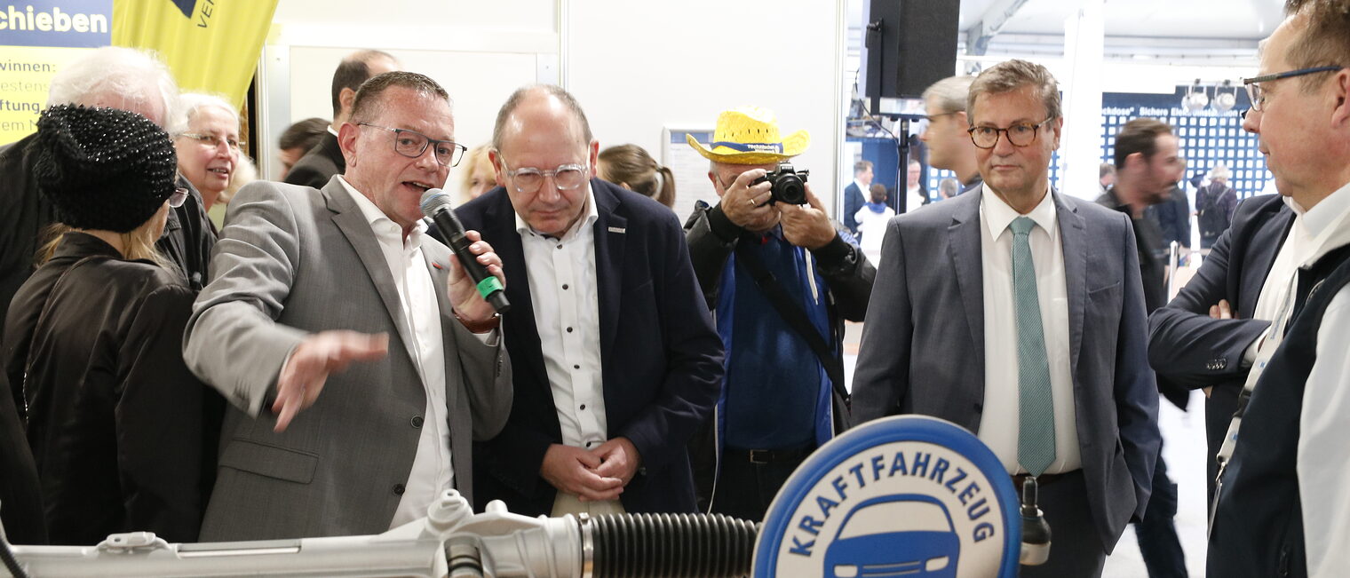 Handwerkskammerpräsident erklärt mit Mikrophon in der Hand einem anderen Mann einen Motor, umrundet von vielen anderen Menschen