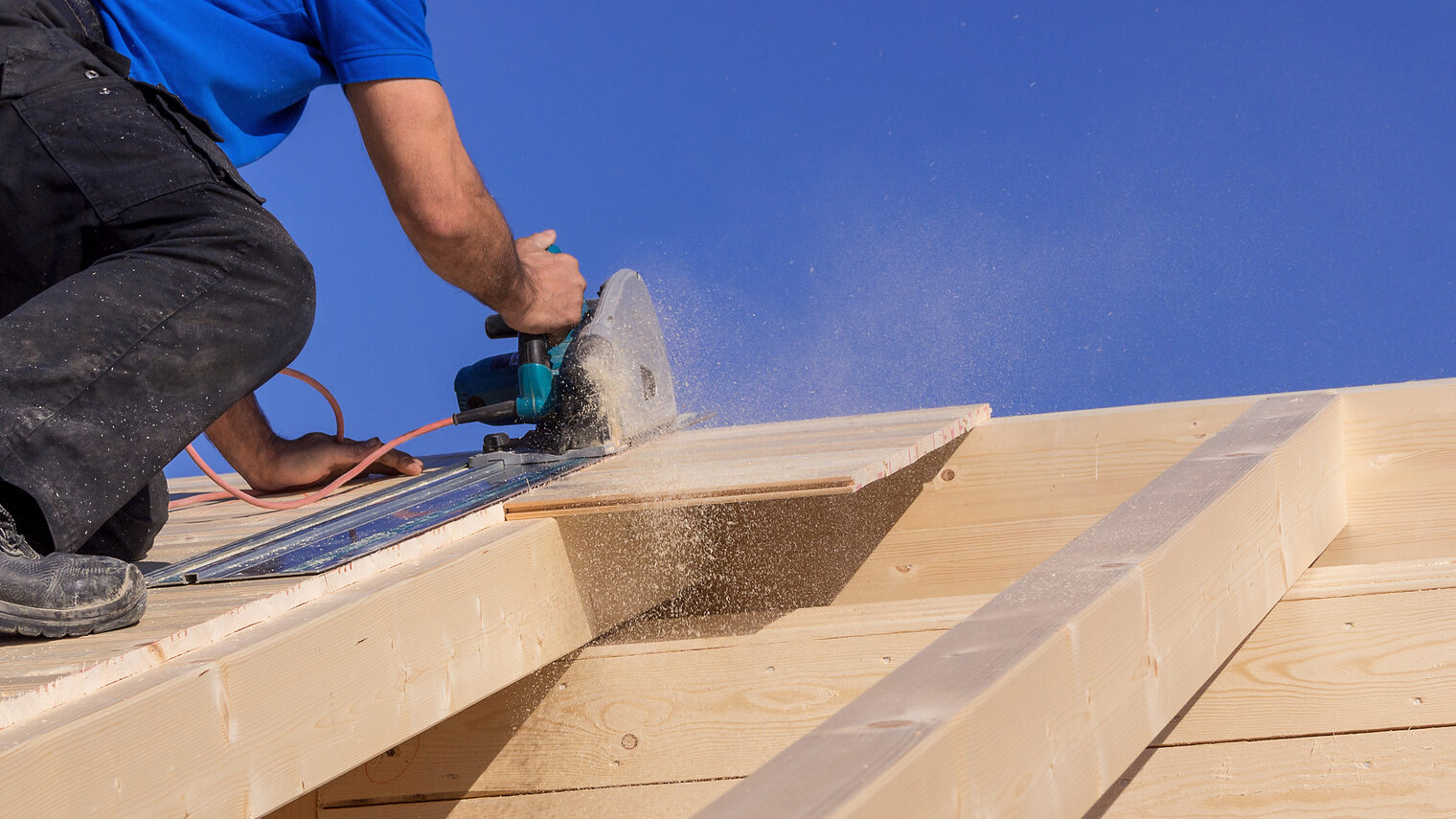 Handwerker sägt auf einem Dach mit Kreissäge bei Sonnenschein, Holzspäne fliegen